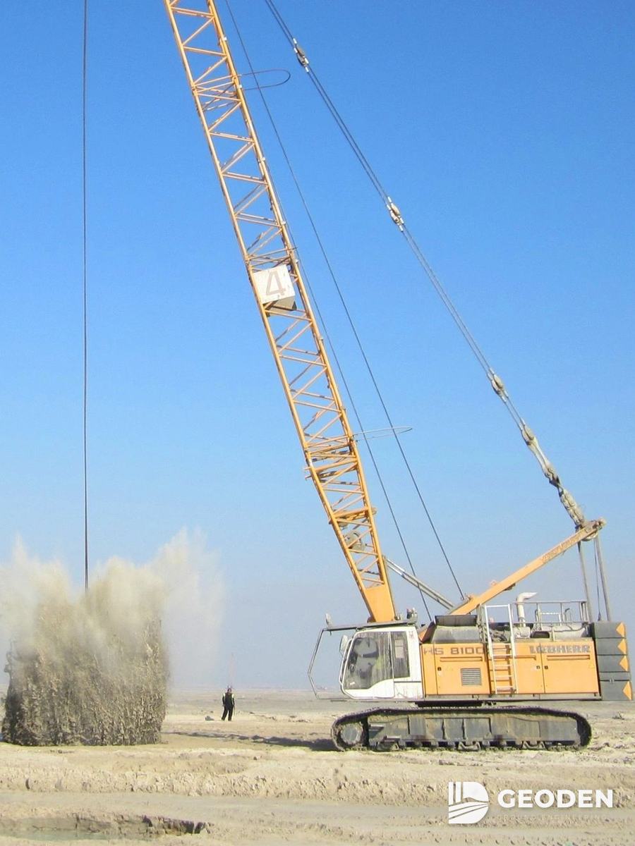 Crawler crane dropping a pounder during dynamic compaction, with a dust cloud at the impact point